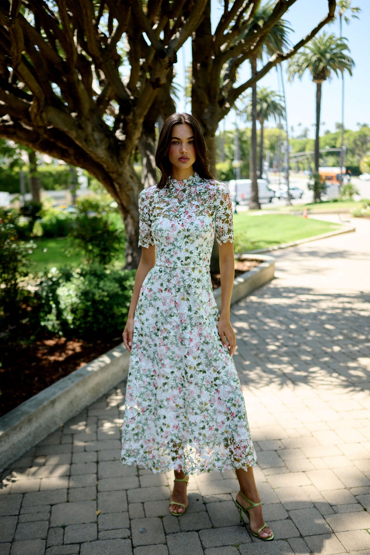 Monique Lhuillier Fall 2026 Silk White/Multi Ivy Bloom Printed Lace Short sleeve day dress with high neckline - model on stone walkway outside.